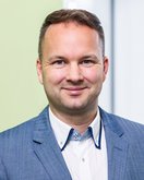 Ein Mann in einem blauen Anzug mit einem weißen Hemd und einem hellen Hintergrund lächelt in die Kamera.