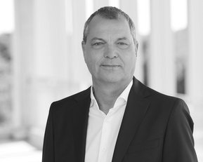 Mann in Anzug mit offenem Hemdkragen, steht vor einer hellen, modernen Architektur. Hintergrund unscharf.