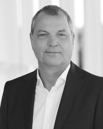 Mann in Anzug mit offenem Hemdkragen, steht vor einer hellen, modernen Architektur. Hintergrund unscharf.