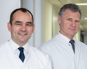 Zwei Ärzte in weißen Kitteln stehen in einem Krankenhausflur und lächeln in die Kamera.