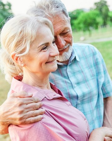 Ein älteres Paar umarmt sich liebevoll im Freien, umgeben von Bäumen und Wiese, während sie einander anlächeln.