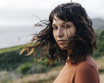 Eine Frau mit lockigem, braunem Haar und Sommersprossen steht im Freien, während der Wind durch ihr Haar weht. Im Hintergrund sind sanfte Hügel und das Meer sichtbar.