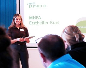 Eine Frau präsentiert einen MHFA Ersthelfer-Kurs vor einer Gruppe von Teilnehmern. Auf der Leinwand sind Kursdetails sichtbar.