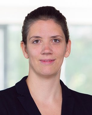 Eine Frau mit einem schwarzen Blazer steht mit verschränkten Armen in einem hellen, modernen Raum. Sie blickt direkt in die Kamera und hat eine neutrale Miene. Der Hintergrund ist unscharf und minimalistisch.