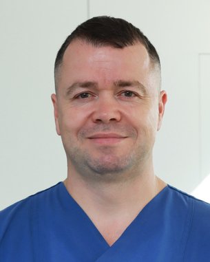 Ein Mann in blauer medizinischer Kleidung steht vor einer hellen Wand und lächelt leicht.