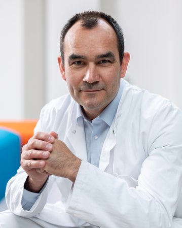 Ein Mann in einem weißen Kittel sitzt mit gefalteten Händen. Er hat kurzes, dunkles Haar und schaut direkt in die Kamera.