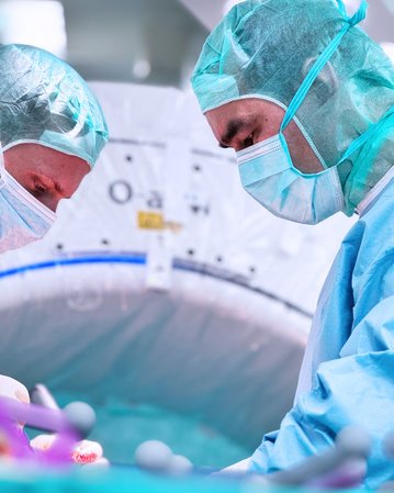 Zwei Chirurgen in OP-Kleidung führen einen Eingriff durch, während sie sich auf den Patienten konzentrieren. Beide tragen Mundschutz und Hauben, um sterile Bedingungen aufrechtzuerhalten.