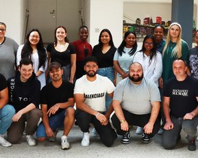 Eine Gruppe von 20 Personen steht zusammen in einem Innenraum. Die Teilnehmer sind vielfältig in Geschlecht und ethnischer Zugehörigkeit, einige tragen lässige Kleidung. Im Hintergrund sind Regale sichtbar.