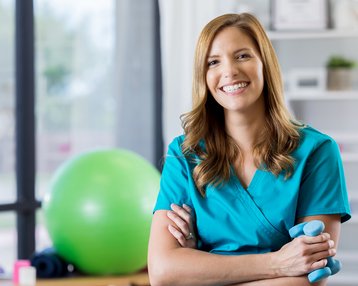Eine lächelnde Frau in blauer Arbeitskleidung steht mit verschränkten Armen in einem Fitnessraum, im Hintergrund ein grüner Gymnastikball.