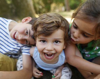 Drei Kinder posieren fröhlich, eines lächelt in die Kamera, während die anderen sich umarmen. Sie sind im Freien, umgeben von Natur.