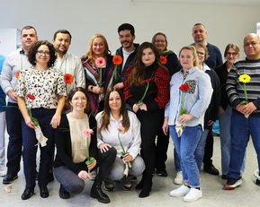 Gruppe von 16 Personen, die Blumen halten, posiert in einem Raum. Ein Flipchart im Hintergrund zeigt den Titel "NIK" und das Datum.