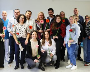 Gruppe von 16 Personen, die Blumen halten, posiert in einem Raum. Ein Flipchart im Hintergrund zeigt den Titel "NIK" und das Datum.