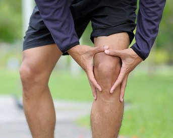 Ein Mann hält sich schmerzhaft das Knie, während er auf einem Gehweg im Freien steht. Er trägt Sportkleidung und sieht betroffen aus.
