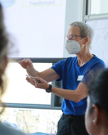 Eine Person mit Brille und Maske erklärt etwas vor einer Gruppe von Zuhörern, während ein Flipchart im Hintergrund steht.