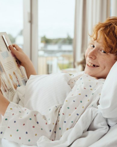 Ein Junge mit roten Haaren liegt im Bett und liest ein Buch. Er trägt ein gepunktetes Nachthemd und lächelt, während er auf die Kamera schaut. Tageslicht strömt durch ein Fenster im Hintergrund.