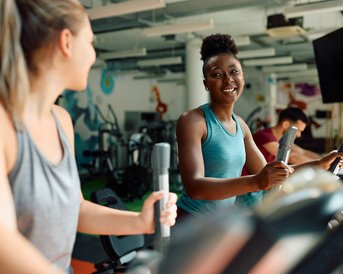 Zwei Frauen trainieren auf Crosstrainern in einem Fitnessstudio, während ein Mann im Hintergrund ebenfalls trainiert.