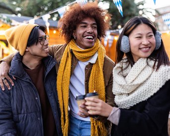 Drei Personen stehen zusammen, lächeln und tragen warme Kleidung. Sie halten Becher in der Hand und stehen vor festlicher Dekoration.