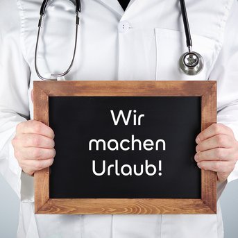 Ein Arzt hält ein Schild mit der Aufschrift "Wir machen Urlaub!" in den Händen.