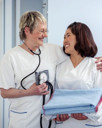 Zwei Krankenschwestern in weißen Uniformen stehen in einem Krankenhauszimmer. Eine hält ein Stethoskop, während die andere einen Stapel Handtücher trägt. Beide lächeln sich an und scheinen eine freundliche Interaktion zu haben.