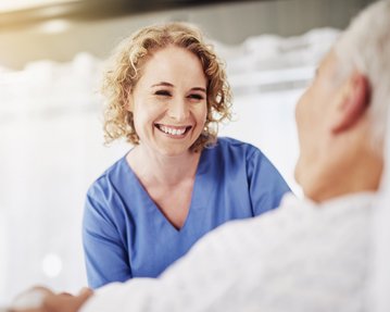 Eine lächelnde Pflegekraft in blauer Uniform spricht mit einem Patienten im Krankenhausbett.