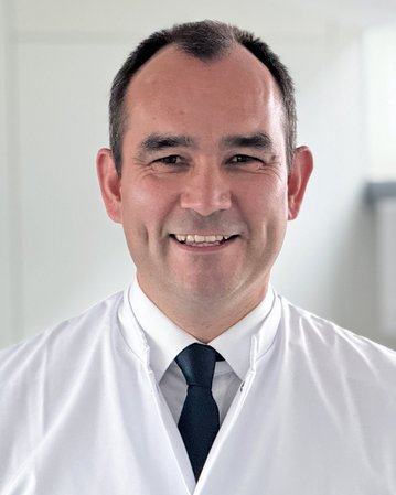 Ein Arzt in einem weißen Kittel steht in einem modernen, hellen Raum und lächelt freundlich. Die Umgebung ist minimalistisch mit großen Fenstern, die Tageslicht hereinlassen.