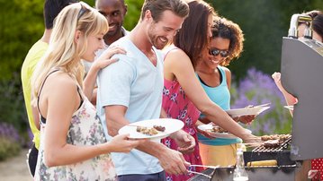 Gruppe von Menschen beim Grillen im Freien, einige halten Teller mit Essen, während andere am Grill stehen und zubereiten.