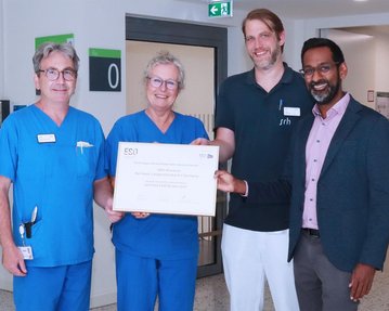 Vier Personen in medizinischer Kleidung halten ein Zertifikat in einem Krankenhausflur. Sie lächeln und stehen nebeneinander.