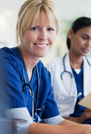 Zwei Krankenschwestern in blauer Arbeitskleidung, eine lächelt und schaut in die Kamera, während die andere Notizen macht.