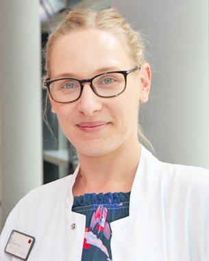 Eine Frau mit Brille trägt einen weißen Kittel und steht lächelnd vor einer neutralen Hintergrundumgebung. Ihr Oberteil hat ein florales Muster in Blau und Rot. Ein Namensschild ist an ihrem Kittel befestigt.