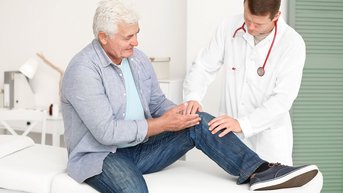 Ein älterer Mann sitzt auf einer Liege, während ein Arzt seine Knieuntersuchung durchführt. Der Arzt trägt einen weißen Kittel und ein Stethoskop.