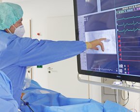 Zwei medizinische Fachkräfte in OP-Kleidung betrachten einen großen Monitor, der Herzfrequenz- und andere Vitaldaten anzeigt. Einer der Fachkräfte zeigt mit dem Finger auf den Bildschirm, während sie Informationen analysieren.