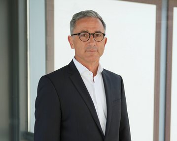 Ein Mann in einem Anzug steht vor einer Glaswand und schaut direkt in die Kamera. Er hat graue Haare und trägt eine Brille.