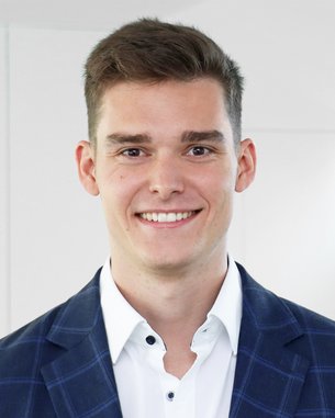 Ein junger Mann mit kurzem, braunem Haar trägt einen blauen Blazer mit Karomuster und lächelt freundlich vor einem hellen Hintergrund.