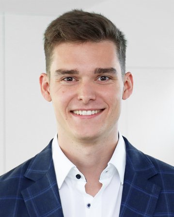 Ein junger Mann mit kurzem, braunem Haar trägt einen blauen Blazer mit Karomuster und lächelt freundlich vor einem hellen Hintergrund.