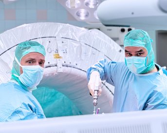 Zwei Chirurgen in OP-Kleidung stehen vor einem medizinischen Gerät und bereiten sich auf einen Eingriff vor.