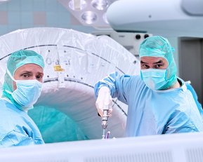 Zwei Chirurgen in blauen OP-Kitteln und grünen Hauben stehen in einem Operationssaal. Einer von ihnen hält ein chirurgisches Instrument, während sie in die Kamera schauen. Im Hintergrund ist ein medizinisches Gerät sichtbar.