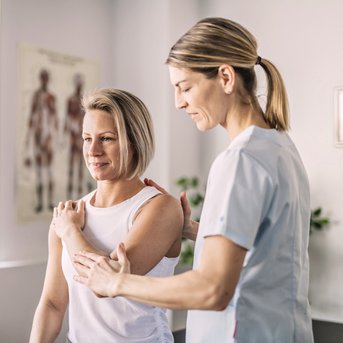 Eine Physiotherapeutin hilft einer Patientin, die ihren Arm bewegt. Die Patientin trägt ein ärmelloses Oberteil und sieht entspannt aus. Im Hintergrund sind anatomische Poster und ein Skelettmodell sichtbar.