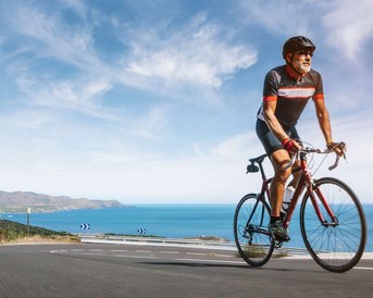 Ein Radfahrer in sportlicher Kleidung fährt auf einer kurvenreichen Straße entlang der Küste mit Blick auf das Meer.