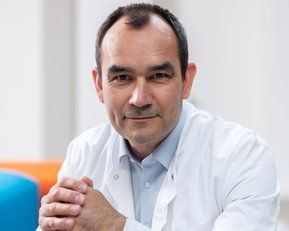 Ein Arzt in einem weißen Kittel sitzt mit gefalteten Händen und lächelt leicht. Der Hintergrund ist unscharf und hell.