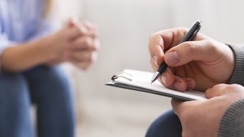 Eine Person hält einen Stift und schreibt in ein Notizbuch. Im Hintergrund sitzt eine andere Person mit gefalteten Händen. Die Szene vermittelt eine Gesprächssituation, möglicherweise in einem Beratungs- oder Therapiekontext.
