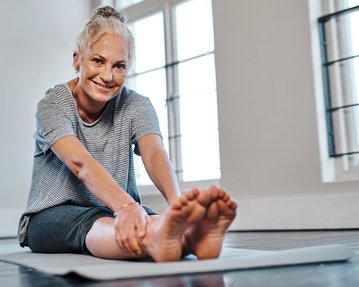 Eine ältere Frau sitzt auf einer Yogamatte und dehnt sich, indem sie ihre Füße erreicht. Sie trägt ein gestreiftes T-Shirt und Shorts.