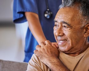 Ein älterer Mann sitzt auf einem Sofa und lächelt, während eine Pflegekraft ihm die Schultern massiert.