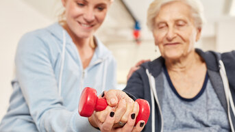 Eine ältere Frau hält ein rotes Hantelgewicht, während eine jüngere Person ihr assistiert. Beide sind in einem Trainingsraum, und die jüngere Person zeigt eine unterstützende Haltung.