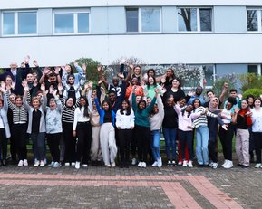 Eine große Gruppe von Menschen steht im Freien und hebt die Hände in die Luft. Sie lächeln und posieren vor einem Gebäude, umgeben von Bäumen und Sträuchern. Die Teilnehmer tragen verschiedene Kleidungsstile.