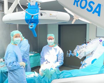 Chirurgisches Team in einem Operationssaal, gekleidet in blaue OP-Kleidung und Masken, mit einem chirurgischen Roboter im Hintergrund.