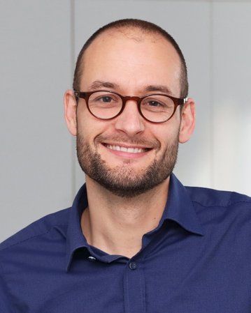 Ein Mann mit Brille und Bart lächelt freundlich in einem hellen Raum, trägt ein blaues Hemd und hat kurze Haare.