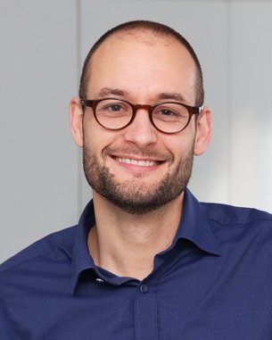 Ein Mann mit Brille und Bart lächelt freundlich in einem hellen Raum, trägt ein blaues Hemd und hat kurze Haare.