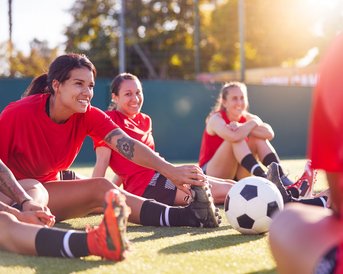 Vier Frauen in roten Trikots dehnen sich auf einem Fußballfeld, während ein Fußball in der Mitte liegt. Sonnenschein im Hintergrund.