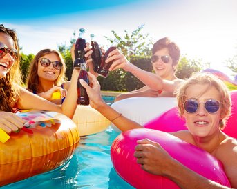 Vier Personen im Pool heben Flaschen mit Getränken an, während sie auf bunten Schwimmreifen sitzen. Sonnenschein und entspannte Atmosphäre.