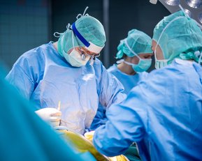 Chirurgisches Team in OP-Kleidung führt eine Operation durch. Drei Personen arbeiten konzentriert an einem Patienten.